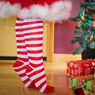 Ms Claus stands on her tip toes to trim a christmas tree, with presents all around.