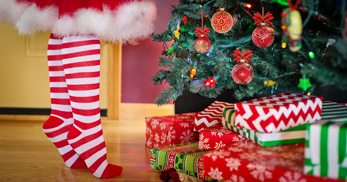 Ms Claus stands on her tip toes to trim a christmas tree, with presents all around.